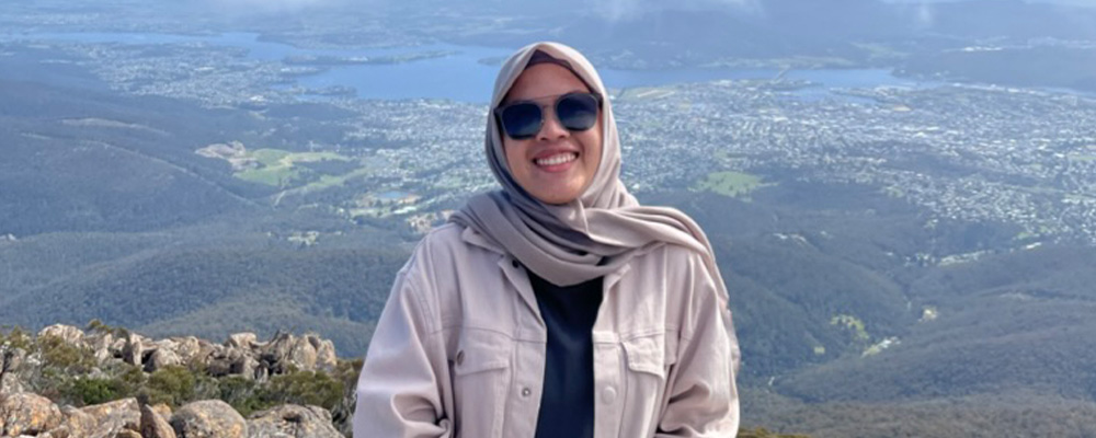 Hasyyati stands, facing the camera and smiling, at the top of a mountain with Canberra below. 