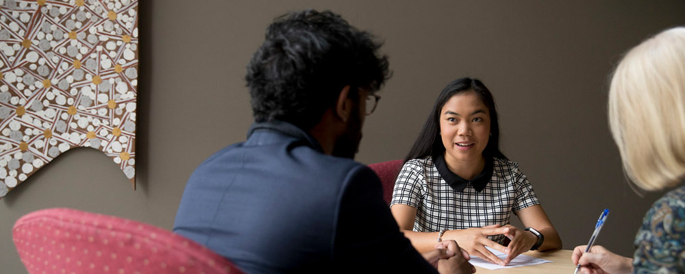 Three students talk to each other around a table and take notes.